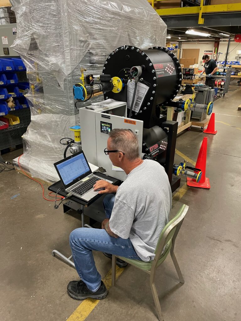 Technician installing industrial burner equipment on a red metal panel at a facility.